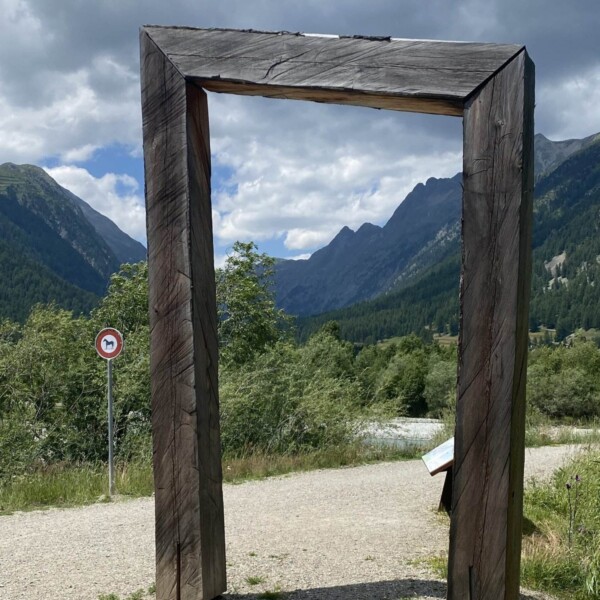 Ein großer rechteckiger Holzrahmen steht auf einem Schotterweg mit Bergen, grünen Bäumen und bewölktem Himmel im Hintergrund. Links vom Rahmen ist ein rundes Verkehrsschild mit rotem Rand zu sehen.