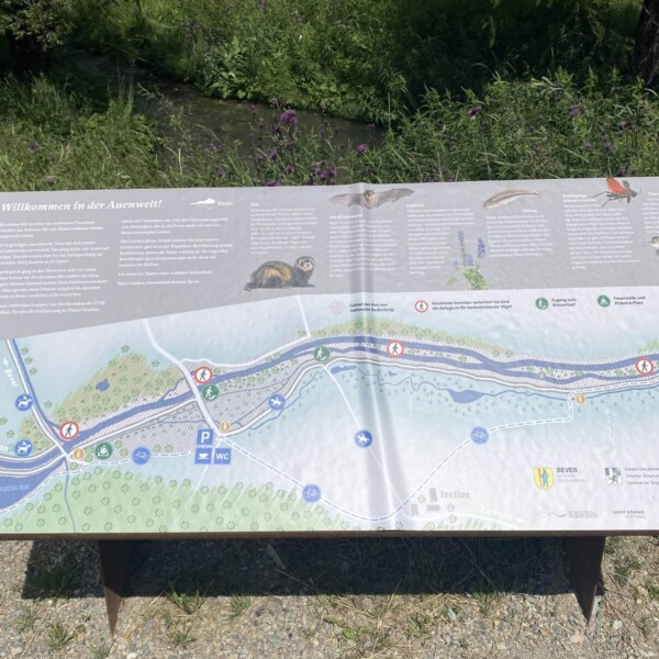 Eine zweisprachige Naturinformationstafel an einem Bach beschreibt die lokale Tierwelt und ihre Lebensräume mit Tierfotos und einer Karte mit Wanderwegen, Sehenswürdigkeiten, Rastplätzen und Parkplätzen in einem Flussuferbereich.