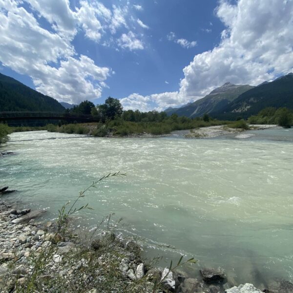 Ein breiter, schnell fließender Fluss mit hellblauem Wasser fließt durch eine felsige, grüne Landschaft. Im Hintergrund sind Berge und Wälder unter einem teilweise bewölkten Himmel zu sehen.