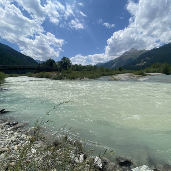 Ein breiter, schnell fließender Fluss mit hellblauem Wasser fließt durch eine felsige, grüne Landschaft. Im Hintergrund sind Berge und Wälder unter einem teilweise bewölkten Himmel zu sehen.