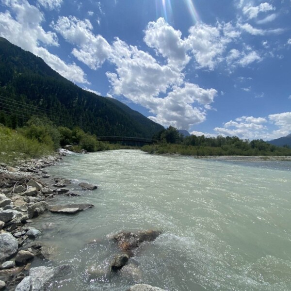 Ein Fluss mit trübem, hellblauem Wasser fließt durch eine felsige Landschaft, gesäumt von grünen Hügeln und Bäumen, unter einem teilweise bewölkten Himmel mit herabströmenden Sonnenstrahlen. Im Hintergrund erheben sich Berge.