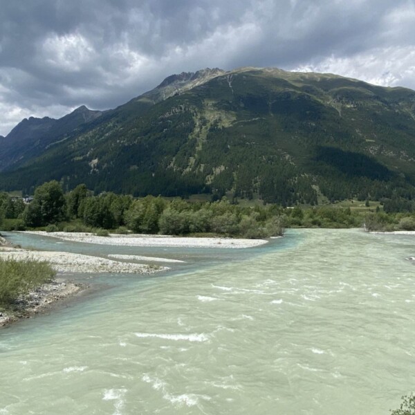 Ein helltürkisfarbener Fluss teilt sich um Kiesinseln, mit üppigen grünen Bäumen und einem bewaldeten Berg im Hintergrund, der sich unter einem bewölkten Himmel erhebt.