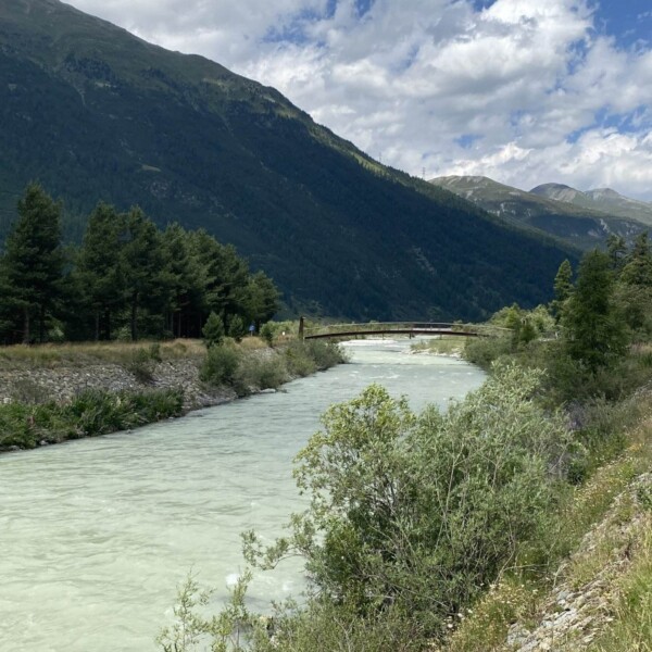 Ein Fluss mit hellblauem Wasser fließt durch ein grünes Tal, umgeben von Bäumen und Büschen. Im Hintergrund erheben sich Berge unter einem teilweise bewölkten Himmel, in der Ferne überquert eine Brücke den Fluss.