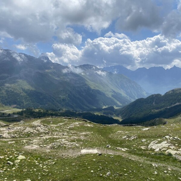Felsige und grasbewachsene Berglandschaft in der Nähe des Berninapasses, mit dem Lago Bianco, der in der Ferne unter einem teilweise bewölkten Himmel schimmert, sanften Hügeln, fernen Gipfeln und Sonnenlicht, das durch die Lücken in den Wolken strömt.