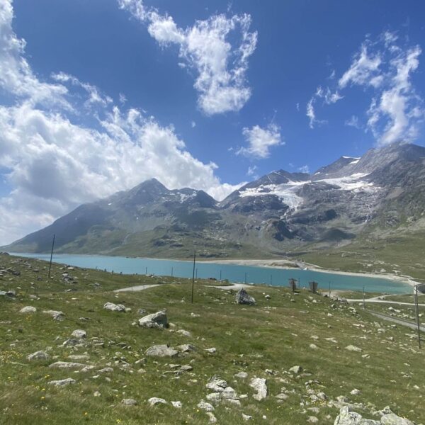 Ein malerischer Blick auf den Lago Bianco, einen türkisfarbenen See auf dem Berninapass, umgeben von Grasfeldern und felsigen Bergen, mit Schneeflecken auf den Gipfeln unter einem teilweise bewölkten blauen Himmel.