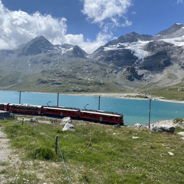 Ein roter Zug fährt am türkisfarbenen Lago Bianco am Berninapass entlang. Im Hintergrund schroffe Berge und schneebedeckte Flächen unter einem leicht bewölkten Himmel. Grünes Gras und Felsen prägen den Vordergrund.