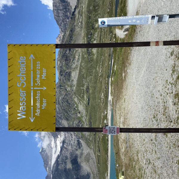 Auf dem Berninapass steht ein gelbes Schild mit der Aufschrift „Wasser Scheide“. Pfeile zeigen links zum „Adriatischen Meer“ und rechts zum „Schwarzen Meer“. In der Nähe sind ein Messmast und der malerische Lago Bianco in der Berglandschaft zu sehen.