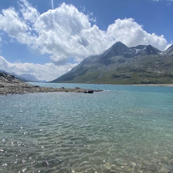 Klarer, türkisfarbener See mit plätscherndem Wasser im Vordergrund, felsiger Uferlinie links und hohen, grünen Bergen des Berninapasses, die sich hinter dem Lago Bianco erheben, ihre schneebedeckten Flecken unter einem teilweise bewölkten blauen Himmel.