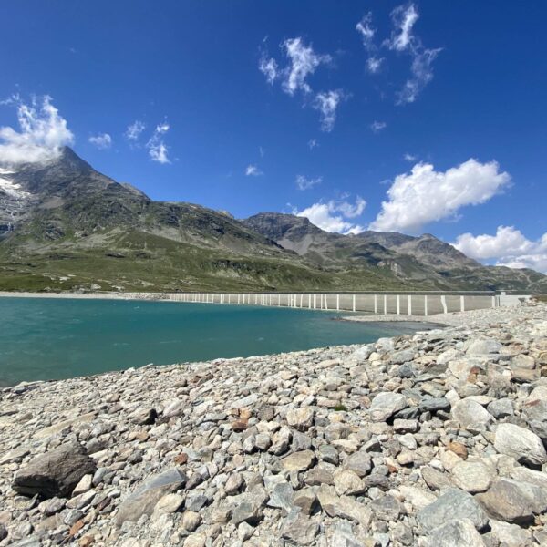Felsiges Ufer am Lago Bianco, einem türkisfarbenen See in der Nähe des Berninapasses, mit einer langen weißen Brücke. Im Hintergrund erheben sich grüne Hügel und schneebedeckte Berge unter einem strahlend blauen Himmel mit vereinzelten Wolken.