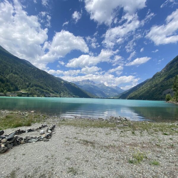Ein ruhiger Bergsee, der Lago di Poschiavo, mit klarem blaugrünem Wasser, umgeben von üppig grünen Hügeln und bewaldeten Bergen unter einem teilweise bewölkten Himmel. Steine und Gras säumen das felsige Ufer im Vordergrund.