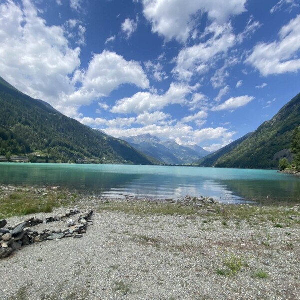 Ein ruhiger Bergsee, der Lago di Poschiavo, mit klarem blaugrünem Wasser, umgeben von üppig grünen Hügeln und bewaldeten Bergen unter einem teilweise bewölkten Himmel. Steine und Gras säumen das felsige Ufer im Vordergrund.