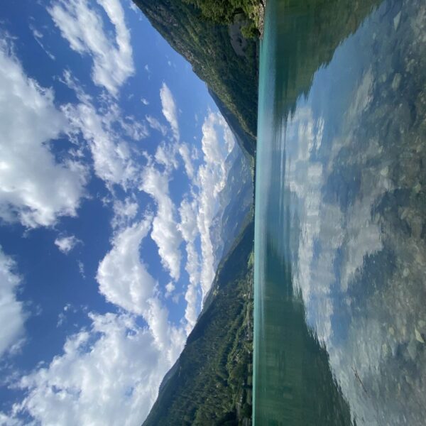Klarer See, umgeben von grünen Bergen, in denen sich der teilweise bewölkte blaue Himmel spiegelt. Die fernen Alpen und Wolken bilden eine ruhige, malerische Landschaft am Lago di Poschiavo. In Ufernähe sind Felsen unter Wasser sichtbar.