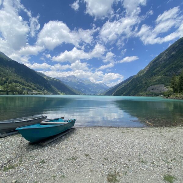 Zwei kleine Ruderboote liegen an einem Kieselstrand am klaren Wasser des Lago di Poschiavo in der Schweiz festgemacht, umgeben von grünen Bergen unter einem leicht bewölkten Himmel. Der See spiegelt den blauen Himmel und die vorbeiziehenden Wolken wider.
