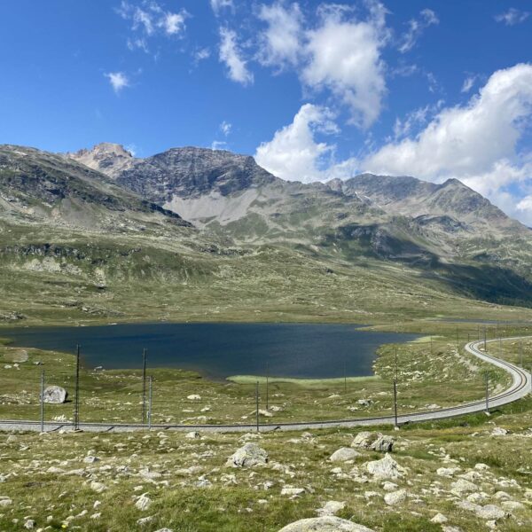 Eine kurvenreiche Straße führt um Lej Nair herum, einen kleinen See in einem grünen Tal entlang des Berninapasses, umgeben von felsigen Hügeln und fernen Bergen unter einem teilweise bewölkten blauen Himmel.