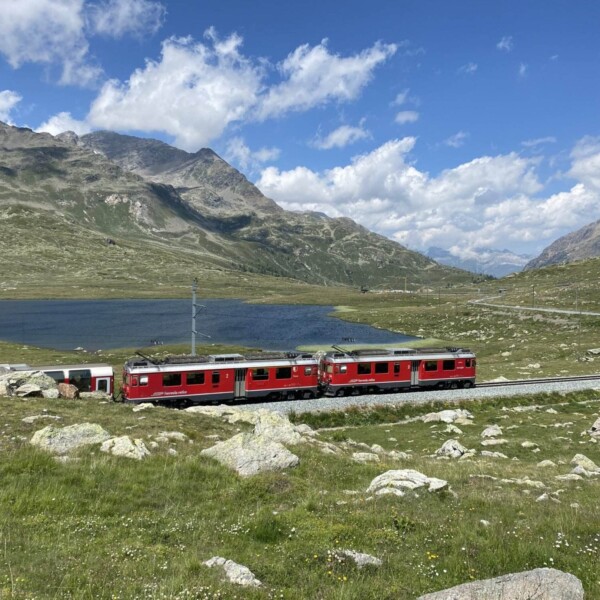 Ein roter Zug fährt durch eine malerische Berglandschaft in der Nähe des Berninapasses mit grünen Hügeln, felsigem Gelände, dem ruhigen See Lej Nair und einem blauen Himmel mit vereinzelten Wolken.