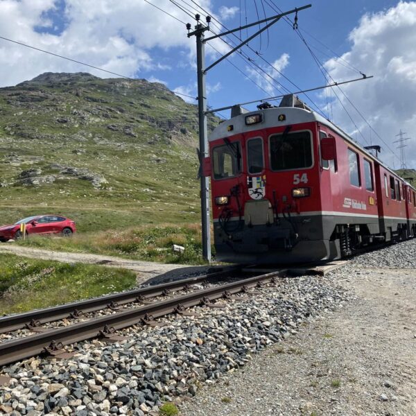 Ein roter Zug fährt auf Gleisen über den Berninapass und schlängelt sich unter leicht bewölktem Himmel an grünen Hügeln und felsigen Hängen vorbei. In der Nähe wartet ein rotes Auto an einem Bahnübergang. Der See Lej Pitschen liegt eingebettet in die Berglandschaft.