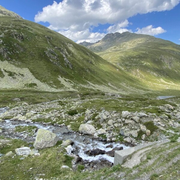 Eine Berglandschaft mit grünen Grashügeln, felsigem Gelände und dem Bach Ova da Radönt, der unter einem teilweise bewölkten blauen Himmel durch das Tal fließt.