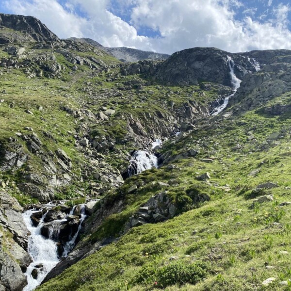 Unter einem teilweise bewölkten blauen Himmel fließt ein Wasserfall die felsigen, grünen Berge von Ova da Radönt hinab, wobei das Sonnenlicht die grasbewachsenen Hänge und das zerklüftete Gelände erhellt.