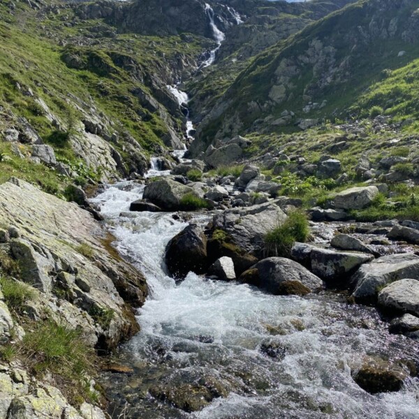 Ein klarer Gebirgsbach, die Ova da Radönt, fließt unter einem strahlend blauen Himmel mit vereinzelten weißen Wolken über Felsen und Gras einen grünen, felsigen Hang hinunter.