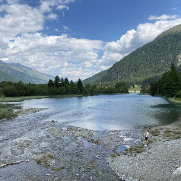 Ein ruhiger See im Val Champagna ist von grünen Hügeln und Wäldern umgeben und steht unter einem teilweise bewölkten Himmel. Zwei Menschen spazieren am felsigen Ufer im Vordergrund entlang, während sich im Hintergrund Berge erheben.