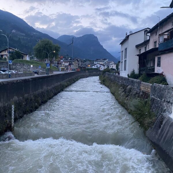 Ein schnell fließender Fluss fließt zwischen Betonmauern durch eine kleine Stadt, auf beiden Seiten sind Häuser zu sehen, im Hintergrund sind unter einem bewölkten Himmel in der Abenddämmerung Berge zu sehen.