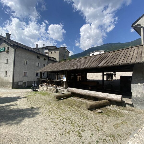 Ein rustikaler Holzunterstand mit Bänken und Tischen steht neben grauen Steingebäuden in einem Bergdorf unter einem teilweise bewölkten Himmel. Im Hintergrund sind grüne Hügel und Berge zu sehen.