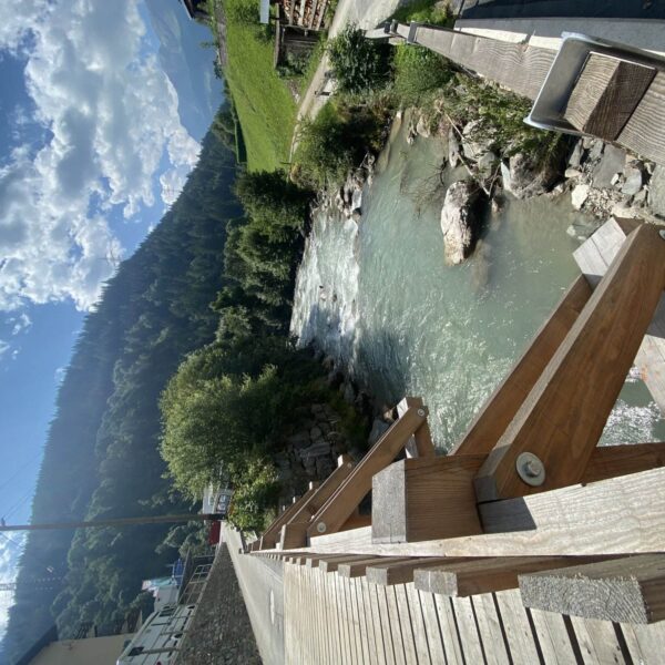 Eine Holzbrücke überquert einen klaren, fließenden Fluss, umgeben von grünen Bäumen, felsigen Ufern und grasbewachsenen Hügeln, mit Bergen und einem teilweise bewölkten Himmel im Hintergrund.