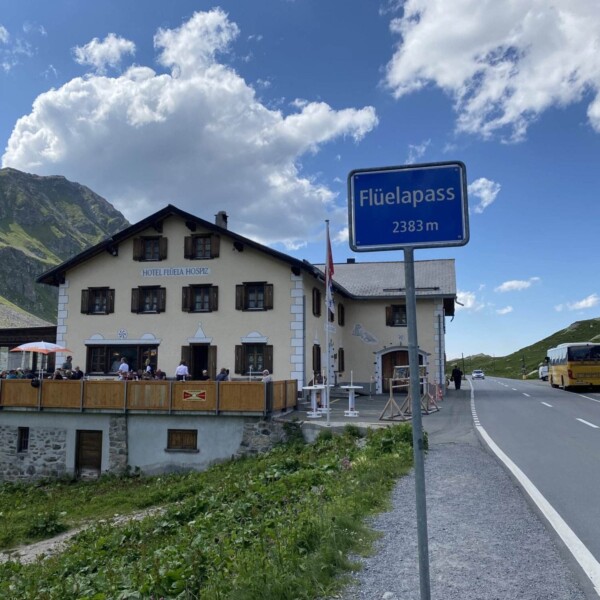 Ein blaues Schild mit der Aufschrift „Flüelapass 2383 m“ steht an einer Bergstraße in der Nähe des Schottensees. Ein cremefarbenes Gebäude und Menschen sitzen auf einer Terrasse. Grüne Hügel und ein teilweise bewölkter Himmel bilden eine malerische Alpenkulisse.