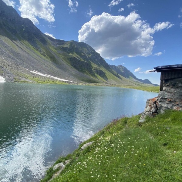 Ein ruhiger Bergsee, der Schottensee, gesäumt von grasbewachsenen Hängen und felsigen Gipfeln unter einem teilweise bewölkten Himmel in der Nähe des Flüelapasses, mit einer kleinen Steinhütte auf der rechten Seite und Spiegelbildern von Wolken und Bergen, die auf der Wasseroberfläche schimmern.