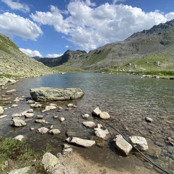 Ein klarer Bergsee, wie der Schwarzsee in der Nähe des Flüelapasses, ist von felsigen Hügeln und grünen Hängen unter einem teilweise bewölkten blauen Himmel umgeben, mit Steinen, die im Vordergrund entlang des Ufers verstreut sind.