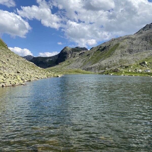 Ein klarer Bergsee, wie der Schwarzsee beim Flüelapass, mit plätscherndem Wasser, umgeben von felsigen und grasbewachsenen Hängen unter einem blauen Himmel mit vereinzelten Wolken.