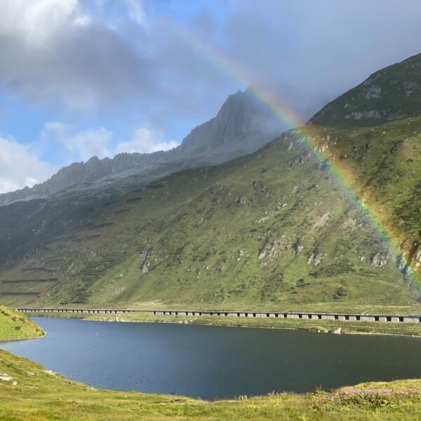 Ein leuchtender Regenbogen spannt sich über eine ruhige Berglandschaft. Im Vordergrund ein ruhiger blauer See, im Hintergrund grüne Hügel, die sich unter leicht bewölktem Himmel zu schroffen Gipfeln erheben. Eine lange Brücke führt am Hang entlang über den See.