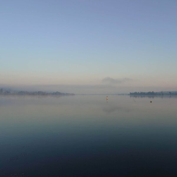 Ein ruhiger, nebliger See im Morgengrauen, dessen stilles Wasser den hellblauen Himmel spiegelt. Schwache Umrisse entfernter Bäume und ein einzelner Mast sind sichtbar und sorgen für eine friedliche und heitere Atmosphäre.