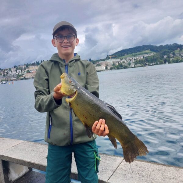 Ein lächelnder Junge in grüner Jacke und Mütze hält einen großen Fisch neben dem Vierwaldstättersee, im Hintergrund sind unter einem bewölkten Himmel eine malerische Schweizer Stadt und Hügel zu sehen.
