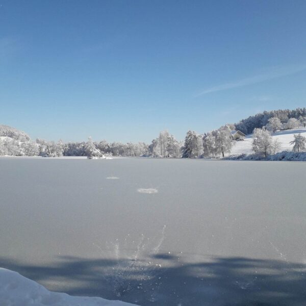 Ein zugefrorener Bettenauer Weiher liegt unter einem strahlend blauen Himmel, umgeben von schneebedeckten Bäumen und Hügeln. Im Vordergrund ist auf der Eisfläche ein Spritzer zu sehen.