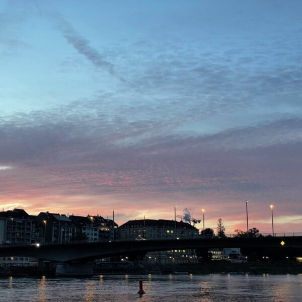 Eine Brücke überquert in der Abenddämmerung einen Fluss. Straßenlaternen leuchten, und am Ufer stehen Gebäude. Der Himmel ist voller rosa, blauer und violetter Wolken, die die Farben des Sonnenuntergangs reflektieren.