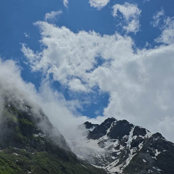 Die Sonne scheint hell über einem teilweise bewölkten blauen Himmel über schneebedeckten Bergen, mit grünen Flecken und aufsteigendem Nebel. Im Vordergrund liegt der ruhige, spiegelnde Schweizer See, der Göscheneralpsee, der zum Wandern in der Nähe einlädt.