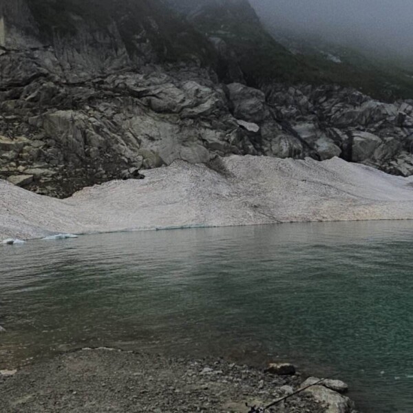 Eine felsige Küste trifft auf das grünliche Wasser des Göscheneralpsees, mit Schnee- und Eisflecken am Fuße der steilen, nebelverhangenen Klippen – eine atemberaubende Schweizer Seenlandschaft, perfekt für Wanderfreunde.