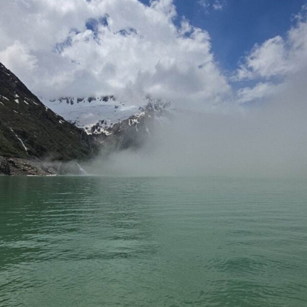 Türkisfarbener Göscheneralpsee mit klarem Wasser im Vordergrund, umgeben von teilweise schnee- und nebelbedeckten Bergen unter einem teilweise bewölkten blauen Himmel – ein atemberaubender Schweizer See, perfekt zum Wandern.