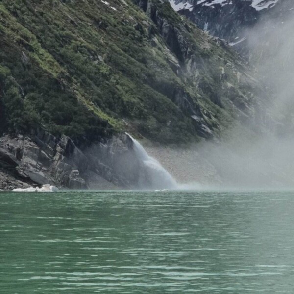 Türkisfarbener Bergsee mit kleinem Wasserfall, der einen grünen, felsigen Berghang in den Schweizer Alpen hinabfließt; im Hintergrund erheben sich schneebedeckte Gipfel unter einem bewölkten Himmel. Nebel schwebt in der Nähe des Wasserfalls.