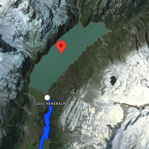 Eine Satellitenkarte zeigt den Göscheneralpsee, einen See in den Schweizer Alpen, umgeben von schneebedeckten Bergen. Eine rote Stecknadel markiert den See, mit einer blauen Wanderroute von der Mautstraße zur Göscheneralp sowie der Beschriftung „Danmahhütte SAC“.