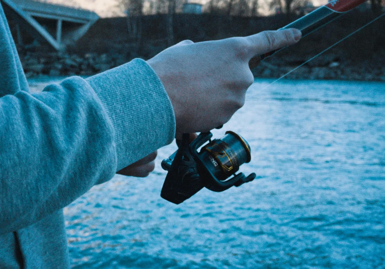 Eine Person in einem grauen Sweatshirt hält eine Angelrute und eine Rolle am Wasser, im unscharfen Hintergrund sind eine Brücke und Bäume zu sehen.