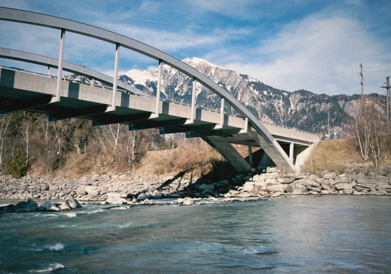 Eine moderne Betonbrücke überquert einen fließenden Fluss, mit felsigen Ufern und schneebedeckten Bergen im Hintergrund unter einem teilweise bewölkten Himmel.