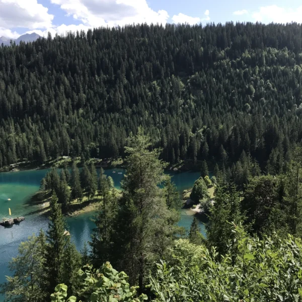 Eine malerische Aussicht auf einen türkisfarbenen See, umgeben von dichten grünen Wäldern und Hügeln unter einem teilweise bewölkten blauen Himmel, wobei das Sonnenlicht die Landschaft erhellt.