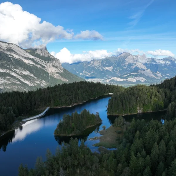 Luftaufnahme eines Waldgebiets rund um einen klaren blauen See mit kleinen Inseln, vor der Kulisse hoher, schroffer Berge unter einem teilweise bewölkten blauen Himmel.