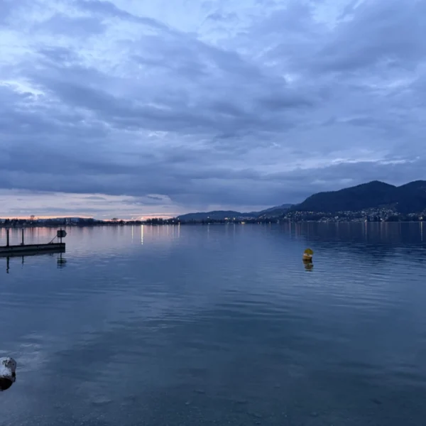 Ein ruhiger See in der Abenddämmerung, in dessen stillem Wasser sich der bewölkte Himmel spiegelt, in der Ferne Hügel am Horizont, links ein Dock, eine schwimmende gelbe Boje und ein schwacher orangefarbener Schein der untergehenden Sonne.