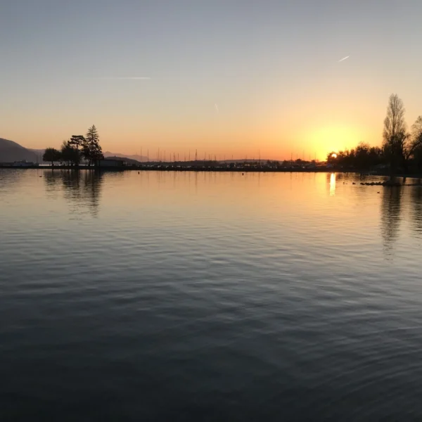 Ruhiger See bei Sonnenuntergang, die Sonne steht tief am Horizont, die Silhouetten von Bäumen, Hügel in der Ferne und eine Reihe von Segelbooten, die sich im stillen Wasser spiegeln und sich vor dem orangefarbenen und blauen Himmel abzeichnen.
