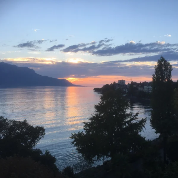 Sonnenuntergang über dem Genfersee (Lac Léman) mit Bergen und Gebäuden am Ufer.