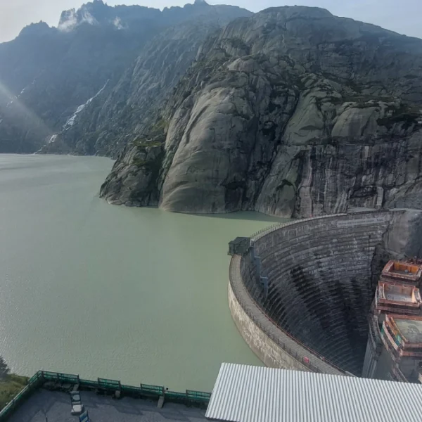Ein großer Damm schlängelt sich zwischen felsigen Bergen an einem See entlang. In der Nähe stehen ein roter Kran und Baumaschinen. Das Sonnenlicht spiegelt sich im Wasser, und im Hintergrund erheben sich steile Klippen unter einem teilweise bewölkten Himmel.