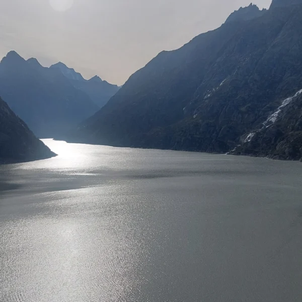 Ein ruhiger See erstreckt sich zwischen steilen, felsigen Bergen unter einem dunstigen Himmel, das Sonnenlicht wird vom Wasser reflektiert und die fernen Gipfel verschwinden im Hintergrund.